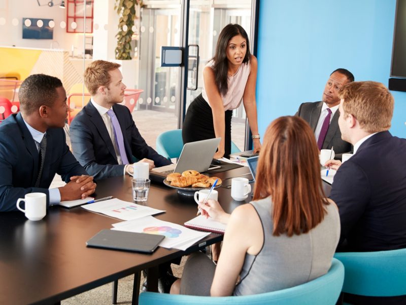 Female manager stands addressing colleagues in meeting room